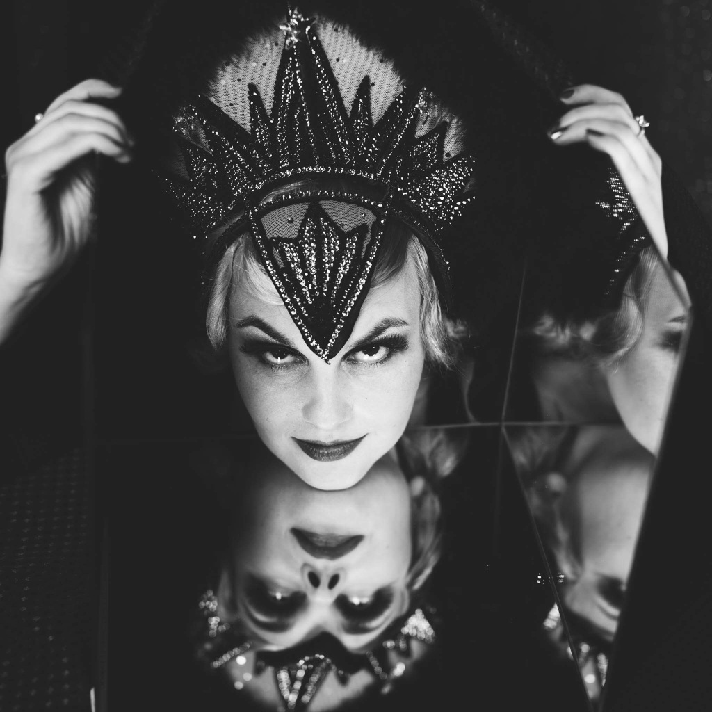 In black and white, a cabaret performer with striking eyebrows and a glamorous crown looks knowingly at the camera. Their face is reflected in a mirror below.