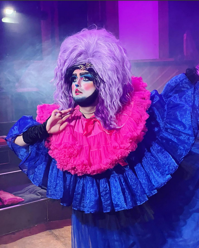 In glorious blue and pink, a queen in a white-painted face and flowing wig poses in forward motion, looking fearfully (or hopefully?) off camera.