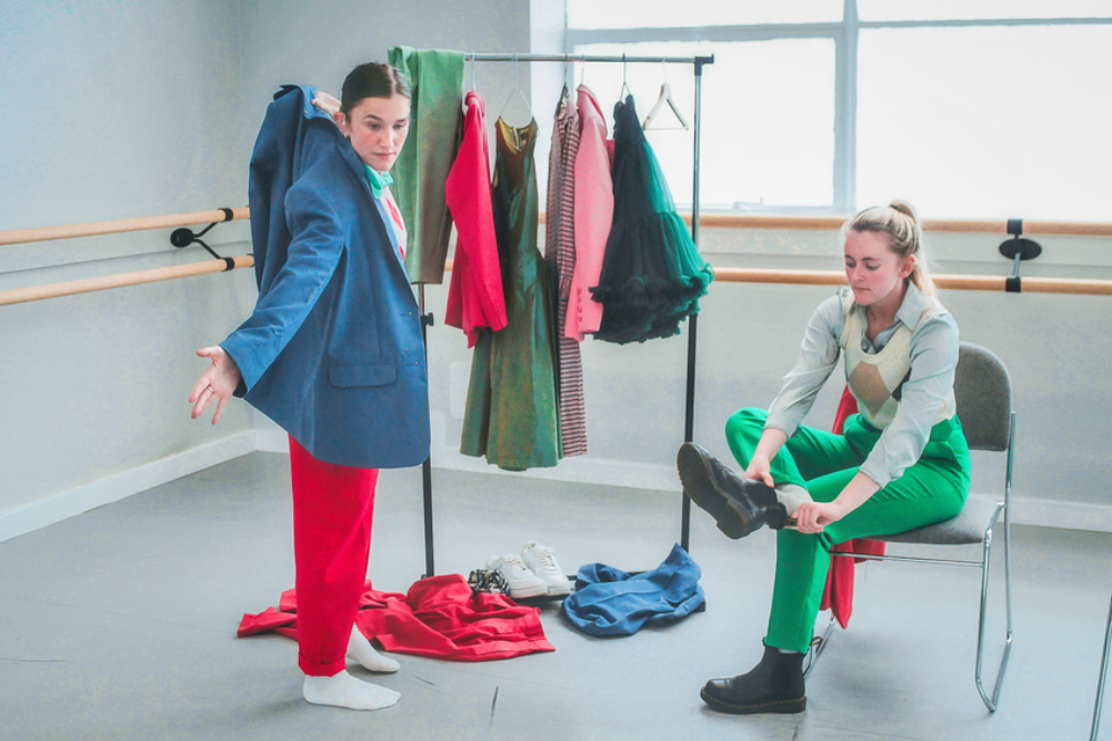 In a well-lit dance studio, two people, one sitting and one standing, try on brightly coloured clothes from a clothes rack.