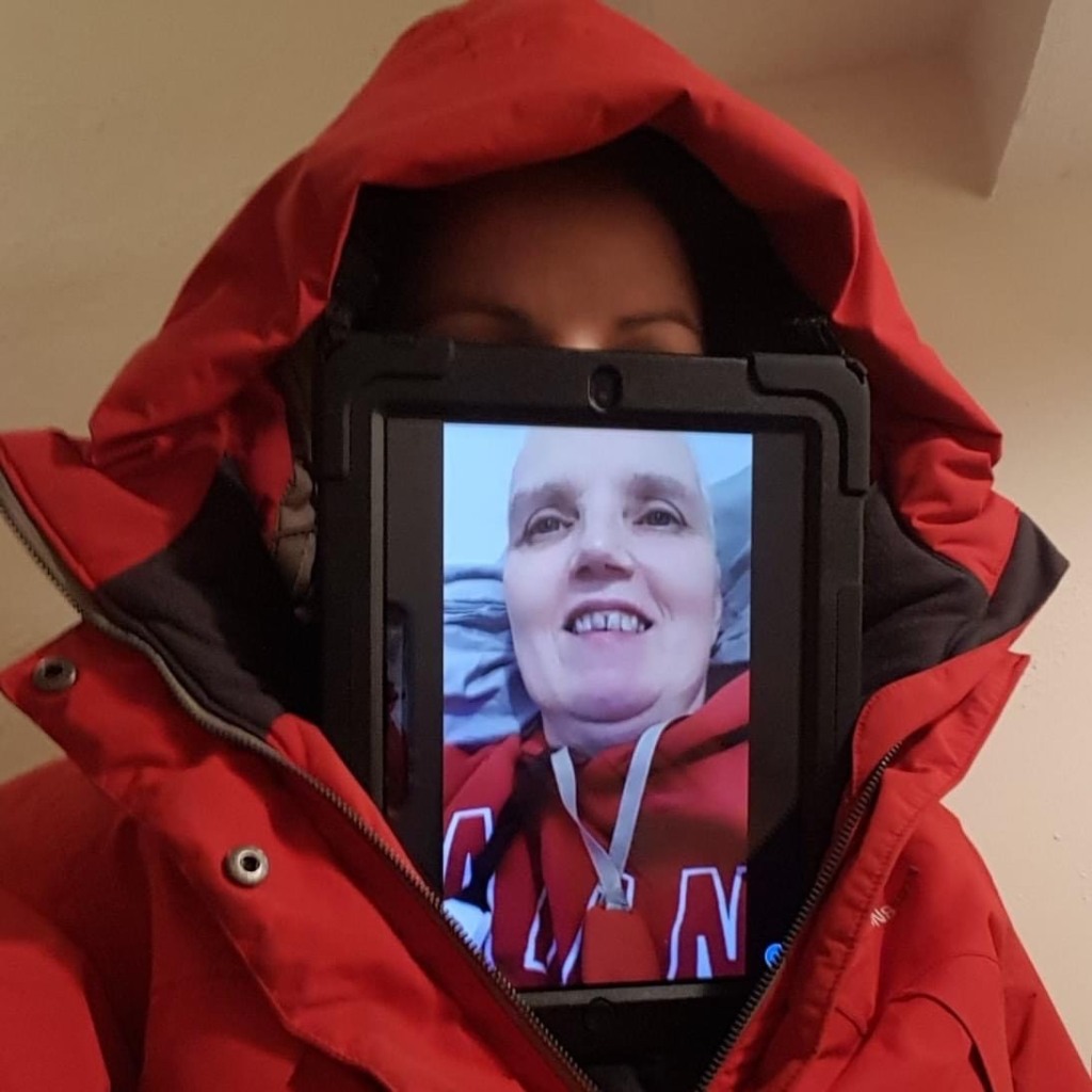 Wearing a large red jacket, the person filling the screen has their face obscured by a large electronic tablet, which features the face of someone reclining in bed.